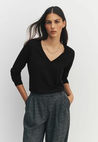 Schwarzer V-Ausschnitt-Pullover mit langen Ärmeln und figurbetontem Design, kombiniert mit grauen strukturierten Hosen mit plissiertem Vorderteil und seitlichen Taschen.