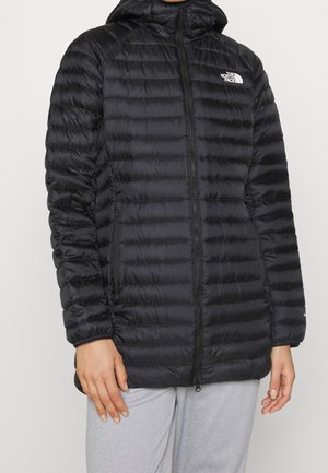 Personne portant une veste matelassée noire à capuche The North Face, fermée par une fermeture éclair, associée à un pantalon gris clair, debout devant un fond uni.