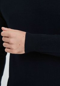 Pull en maille noir avec des poignets côtelés, mettant en valeur une texture lisse et une coupe ajustée. Focus sur la main gauche reposant près de la manche.