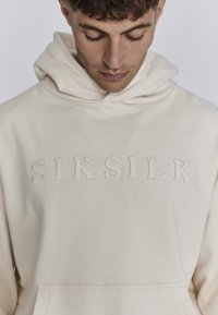 Crèmekleurige hoodie van zacht stof met een voorzak in kangoeroestijl en de ingedrukte tekst "SIKSILK" op de borst.