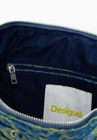 Kék szövet táska belseje ezüst cipzárral, belső cipzáros zsebbel és fehér címkével, rajta sárga "Desigual" felirattal.
