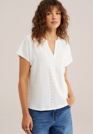 Femme aux cheveux bouclés portant un chemisier blanc à manches courtes avec des broderies et un jean bleu, debout avec une main dans la poche, sur un fond uni.