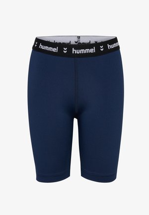 Kind mit verschränkten Armen, trägt marineblaue Sportbekleidung, weiße Socken und blaue Turnschuhe, steht selbstbewusst vor einfachem Hintergrund.
