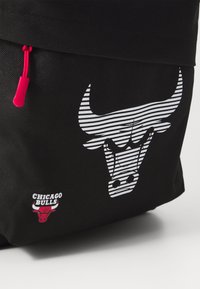 Zwarte rugzak met een wit gestreept logo van de Chicago Bulls, een gekleurde ritslip in het roze en een klein rood-wit merklabel.