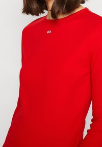 Donna che indossa un top rosso attillato a maniche lunghe con un piccolo logo rettangolare centrato sotto il collo.