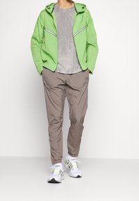 Veste vert clair avec capuche et deux poches zippées à l'avant, associée à une chemise grise texturée et un pantalon beige ample. Baskets blanches avec des accents néon.