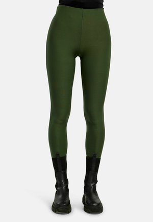 Personne portant des leggings moulants vert olive associés à des bottes en cuir noires épaisses, debout devant un fond uni.