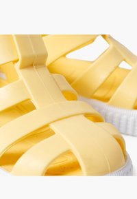 Kollane plastmassist sandaalid, millel on rihma disain, avatud küljed ja tekstureeritud tald valgete aktsentidega.