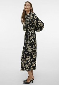 Robe noire longue avec des motifs floraux beiges, col boutonné, poignets élastiques et ceinture assortie. Portée avec des chaussures à talons noires.