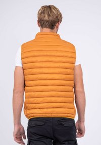 Gilet rembourré orange avec des panneaux matelassés horizontaux, un col haut et un matériau léger. Porté par-dessus une chemise blanche et associé à un pantalon sombre.