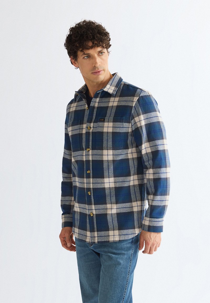 Homme portant une chemise à carreaux bleue et blanche à boutons et un jean bleu, debout et regardant sur le côté sur un fond uni.