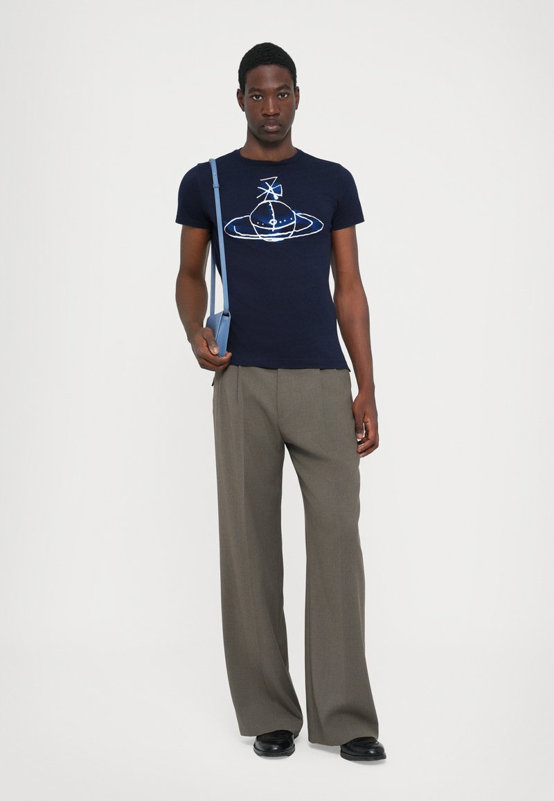 Marineblaues T-Shirt mit weißem Grafikdesign, kombiniert mit hellbraunen weiten Hosen. Das Model hält eine hellblaue Umhängetasche.