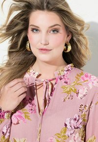 Rosa Bluse mit Blumenmuster, Rüschenausschnitt, Bindebandakzent und langen Ärmeln, die einen leichten Schimmer und mehrfarbige Blumenmuster aufweisen. Goldene Creolen.