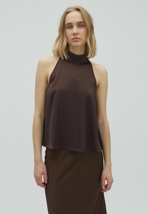 Młoda kobieta o blond włosach do ramion, ubrana w bezrękawnik ciemnobrązowy top z wysokim kołnierzem i pasującą spódnicę, stoi na jednolitym tle.