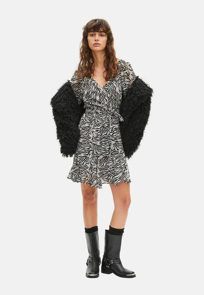 Mujer con vestido de estampado de cebra, una chaqueta negra texturizada colocada sobre los hombros y botas negras hasta la mitad de la pantorrilla con detalles metálicos.