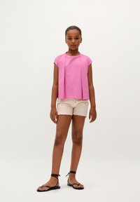 Pink short-sleeve top with a front drape design, paired with beige denim shorts with frayed edges and black flat sandals.