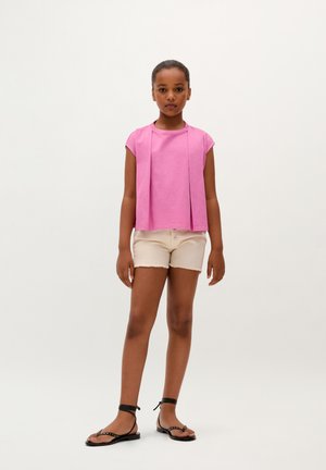Pink short-sleeve top with a front drape design, paired with beige denim shorts with frayed edges and black flat sandals.