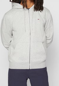 Sudadera gris con cremallera y capucha con cordón, confeccionada en tela suave. Cuenta con bolsillos delanteros y un pequeño logo en el pecho.