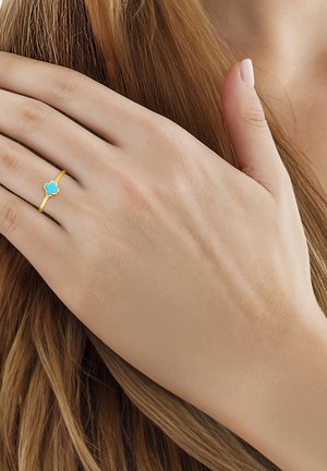 Mano femenina con un anillo de oro que presenta un pequeño trébol de cuatro hojas de turquesa, descansando sobre mechones de cabello castaño claro.