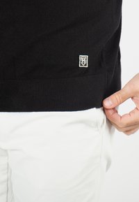 Pull noir en tissu doux, avec un ourlet côtelé et un petit logo brodé en blanc. Associé à un pantalon blanc.