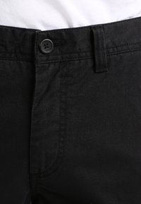Pantalons en tissu noir avec une fermeture à bouton standard et une fermeture éclair, possédant une texture lisse et de subtils détails de couture le long des coutures.
