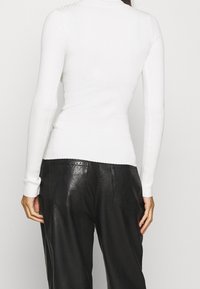 Vrouw met een wit geribd shirt met lange mouwen en zwarte nep-lederen broek, gezien van achteren met ontspannen handen langs haar lichaam.