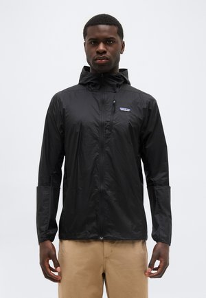 Ung mand iført en sort hættetrøje med lynlås og beige bukser, stående med armene afslappet mod en ensartet grå baggrund.