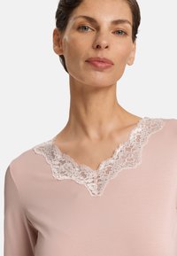 Top rosa claro con escote en V de encaje. La tela es suave y tiene mangas largas, resaltando el delicado detalle de encaje en el cuello.