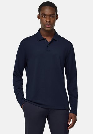 Polo a maniche lunghe di colore blu navy con un colletto a tre bottoni, tessuto morbido e un discreto dettaglio sul fondo, abbinato a pantaloni scuri.