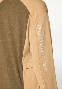 Langsleeve top met een twee-kleurig ontwerp in beige en olijfgroen, gladde textuur en "Peak Performance"-tekst gedrukt op de mouw.