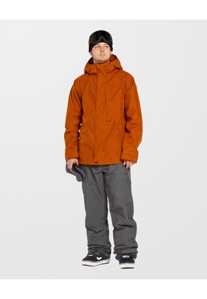 Chaqueta impermeable naranja con capucha, bolsillo frontal con cremallera, pantalones de nieve grises y zapatillas negras, sostenida con guantes grises.