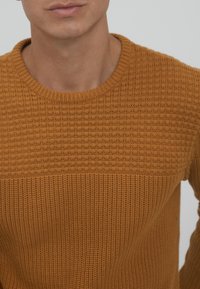 Sennepsfarget strikkegenser med et teksturert mønster, med rund hals og ribbestrikkede mansjetter, vist i overkroppsvinkel.
