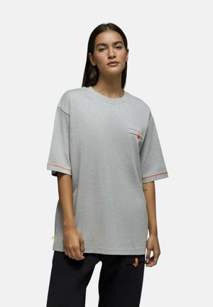 Femme aux longs cheveux foncés portant un t-shirt gris ample à manches courtes avec des accents orange et un pantalon noir sur un fond clair uni.