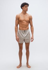 Νεαρός μυώδης άνδρας όρθιος, ξυπόλυτος, φοράει ανοιχτό μπεζ μπόξερ με σχέδιο, μπροστά σε απλό φωτεινό φόντο.