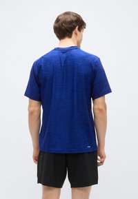 Modra športna majica s kratkimi rokavi in teksturirano površino; ima okrogel izrez in logotip blizu roba. Oblečena s črnimi kratkimi hlačami.