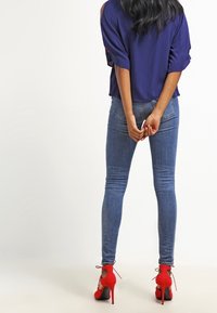 Marineblauw T-shirt met korte mouwen, blauwe skinny jeans en rode hoge hakken met kantdetails. Van achteren weergegeven.