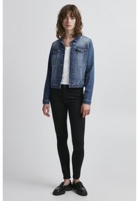 Giacca di jeans di un blu medio con un design corto, dotata di chiusura a bottoni e due tasche sul petto, abbinata a jeans skinny neri e scarpe nere.