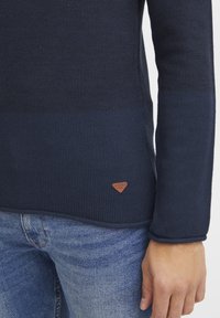 Navyblauwe, gestructureerde trui met lange mouwen, voorzien van een klein, driehoekig leren logo aan de onderkant, gecombineerd met lichtblauwe jeans.