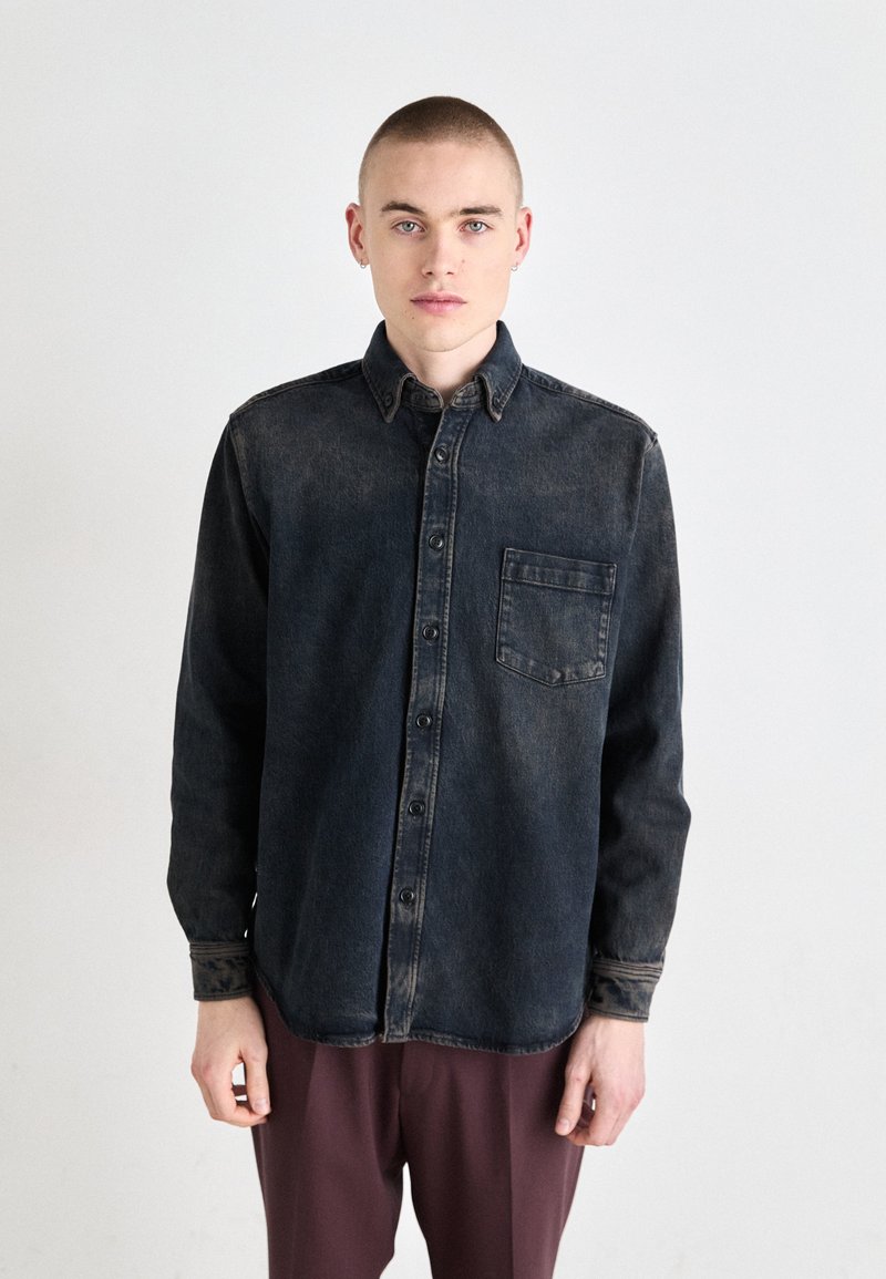 Camicia in denim scuro con bottoni, maniche lunghe, tasca frontale e colletto. Trama liscia con una leggera sfumatura. Indossata con pantaloni bordeaux.