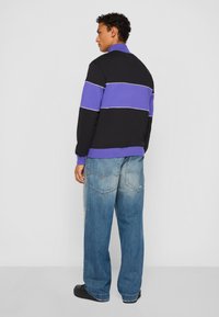 Sort og lilla farveblokkeret sweatshirt med lange ærmer, ribbet krave og hvid aksentstreg, parret med slidte blå vide jeans.