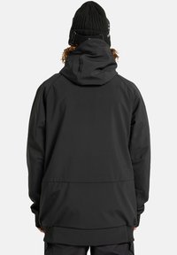 Persona che indossa un giubbotto con cappuccio nero e un berretto di lana nero, vista di spalle su uno sfondo grigio chiaro uniforme.