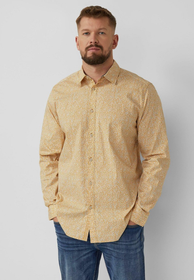 Man met baard, gekleed in een beige overhemd met patroon en lange mouwen en een blauwe spijkerbroek, staand met handen in de zakken tegen een grijze achtergrond.