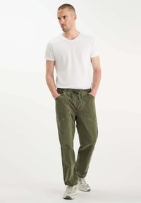 Man in een wit T-shirt met korte mouwen, olijfgroene broek met koord en grote zakken, en beige sneakers, staande met zijn handen in de zakken.