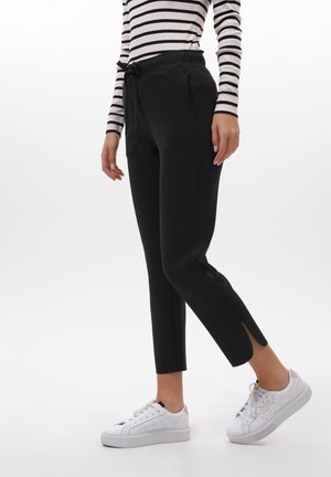 Donna che indossa pantaloni neri affusolati con spacchi alla caviglia, una maglia a maniche lunghe a righe bianche e nere e sneaker bianche su sfondo bianco.