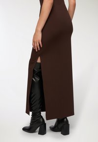 Abito marrone lungo fino al pavimento con uno spacco laterale, che mette in risalto stivali neri sopra il ginocchio e tacchi spessi. Tessuto liscio, design aderente.