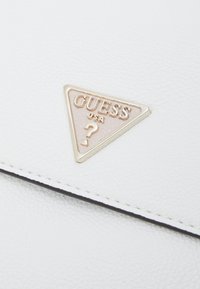 Triangulär metallisk Guess USA-logotyp med ett frågetecken på en strukturerad vit läderyta nära en sydd kant.