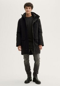 Must hõlmadega must park, millel on lukuga esi, kaks suurel taskut ja elastsed varrukaotsad, kantud tumeda sviitri ja hallide teksade üle.