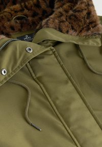 Veste vert olive avec fermeture éclair, boutons-pression en métal, cordons de serrage et un col à capuche doublé en fausse fourrure marron.