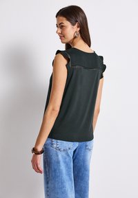 Dunkelgrüne Kurzarmbluse mit Rüschendetail und dekorativen Nähten auf der Rückseite, kombiniert mit hellblauen weitem Jeans.