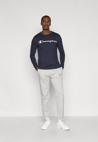 Maglietta a maniche lunghe blu navy con logo "Champion" bianco, abbinata a pantaloni della tuta grigi e sneakers bianche. Abbigliamento sportivo casual, tessuti morbidi.